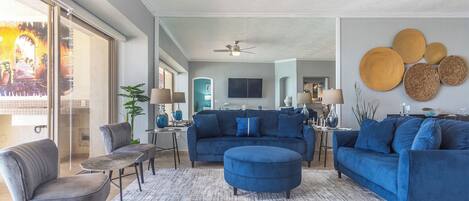Ein modernes Wohnzimmer mit einem blauen Sofagarnitur, einem grauen Sessel und einem Couchtisch. Es gibt einen Deckenventilator, einen großen Spiegel und eine Essecke im Hintergrund.