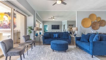 Una sala moderna con un sofá azul, un sillón gris y una mesa de centro. Se observa un ventilador de techo, un espejo grande y un comedor al fondo.