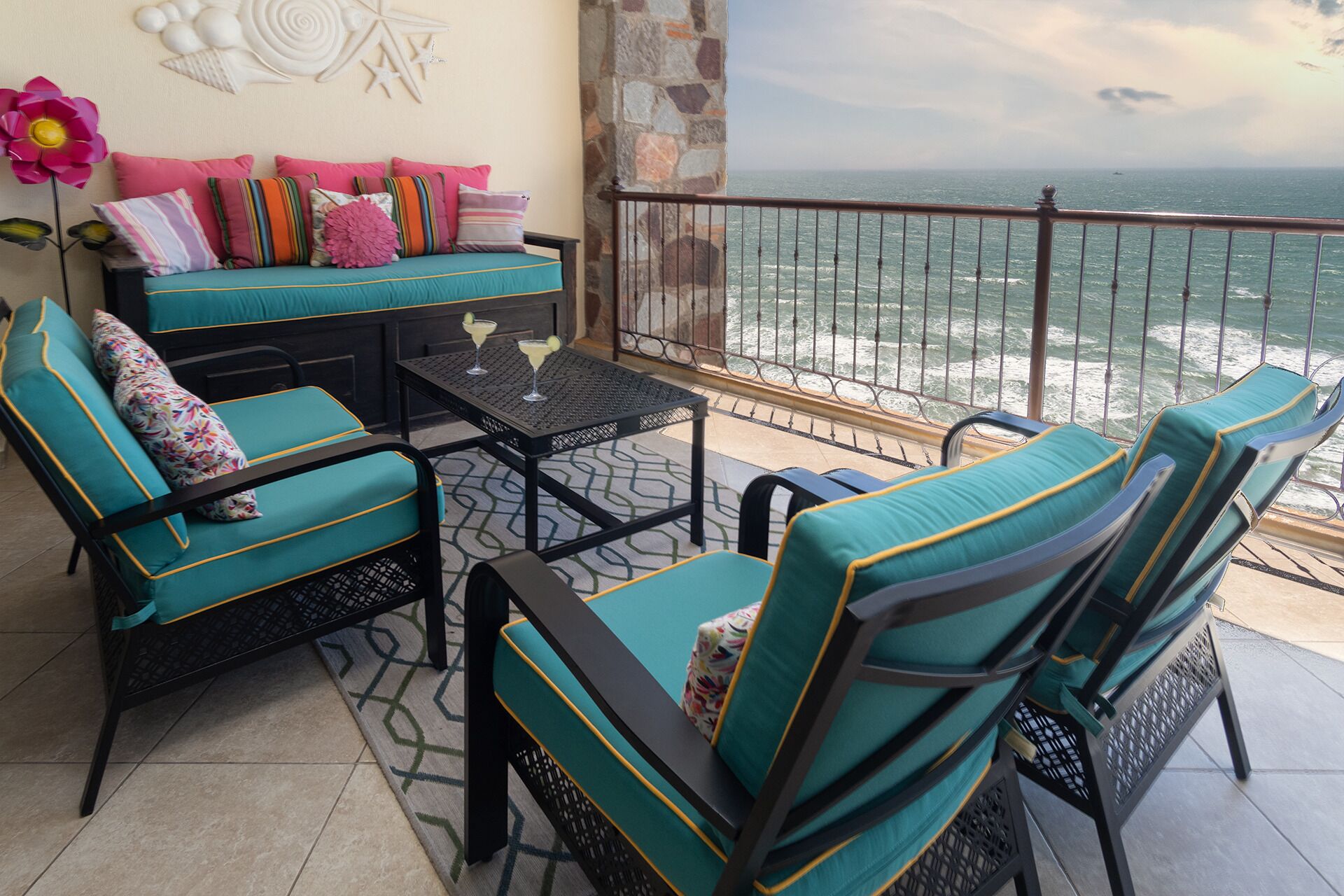 A balcony with turquoise upholstered chairs and a sofa, a small table with drinks, and a view of the ocean.