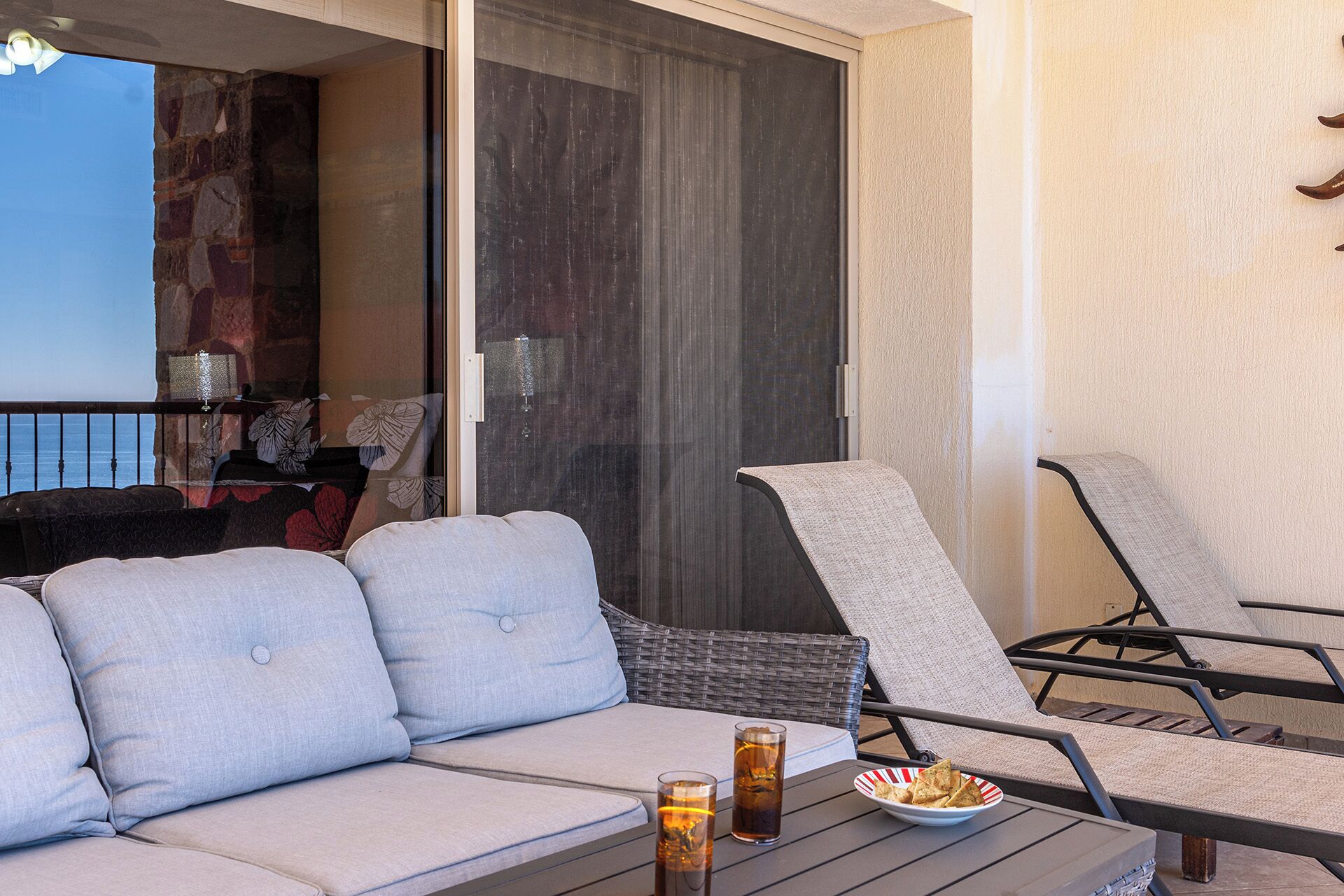 Een balkon met een bank en twee stoelen, een tafel met drankjes en snacks, en uitzicht op de oceaan door een glazen deur.