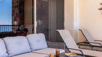 Een balkon met een bank en twee stoelen, een tafel met drankjes en snacks, en uitzicht op de oceaan door een glazen deur.