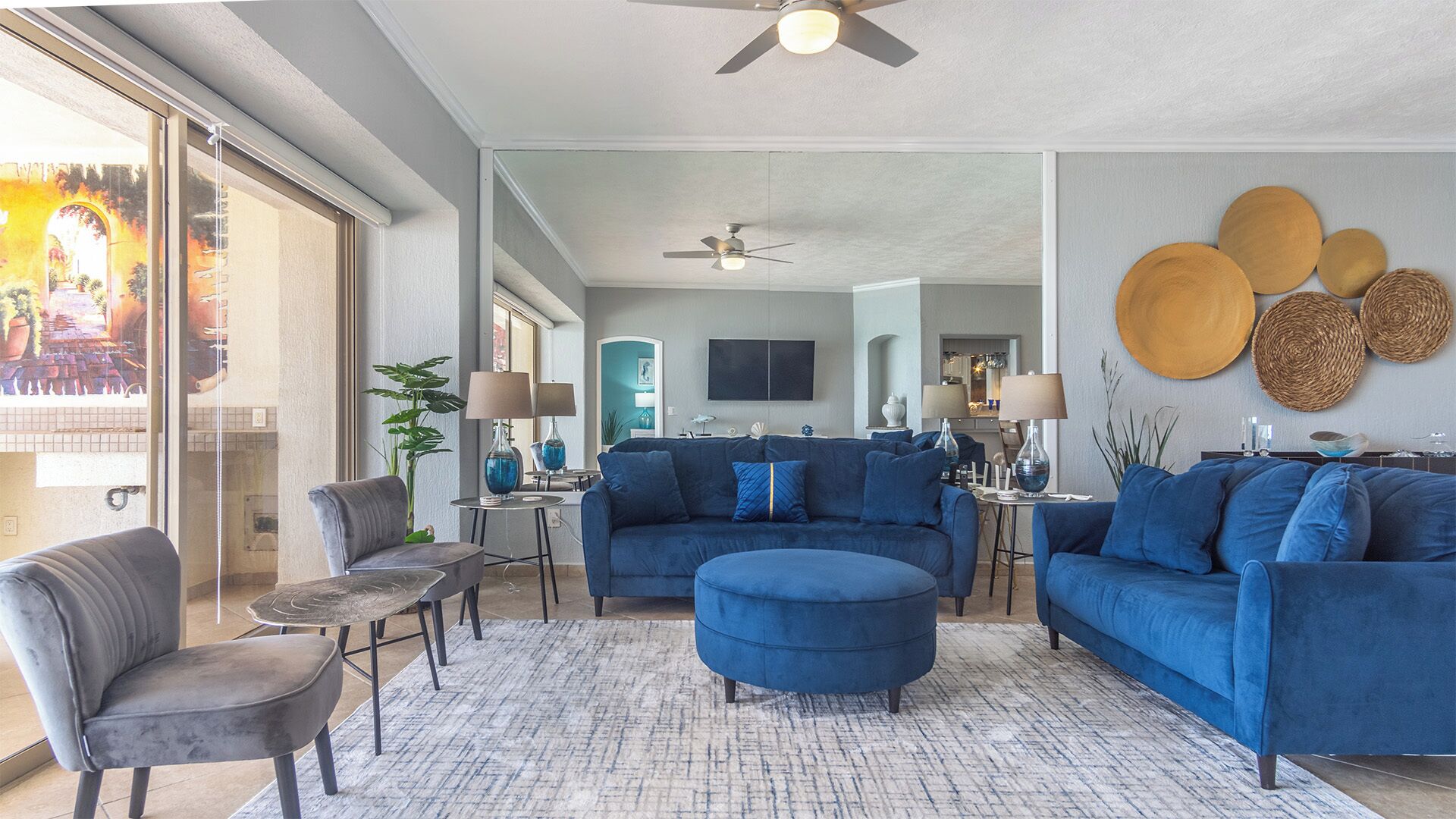 Un salon moderne avec un canapé bleu, un fauteuil gris et une table basse. On peut voir un ventilateur de plafond, un grand miroir et un coin repas à l’arrière-plan.