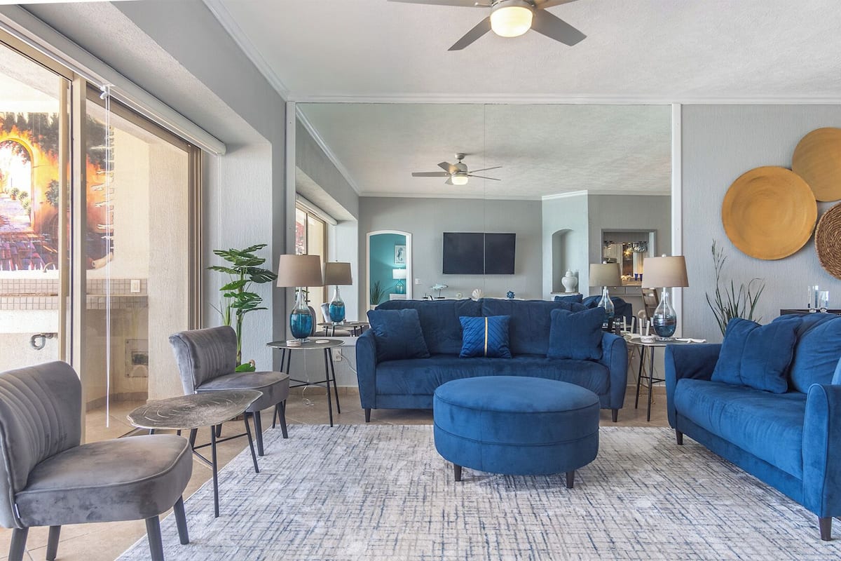 Un salon moderne avec un canapé bleu, un fauteuil gris et une table basse. On peut voir un ventilateur de plafond, un grand miroir et un coin repas à l’arrière-plan.