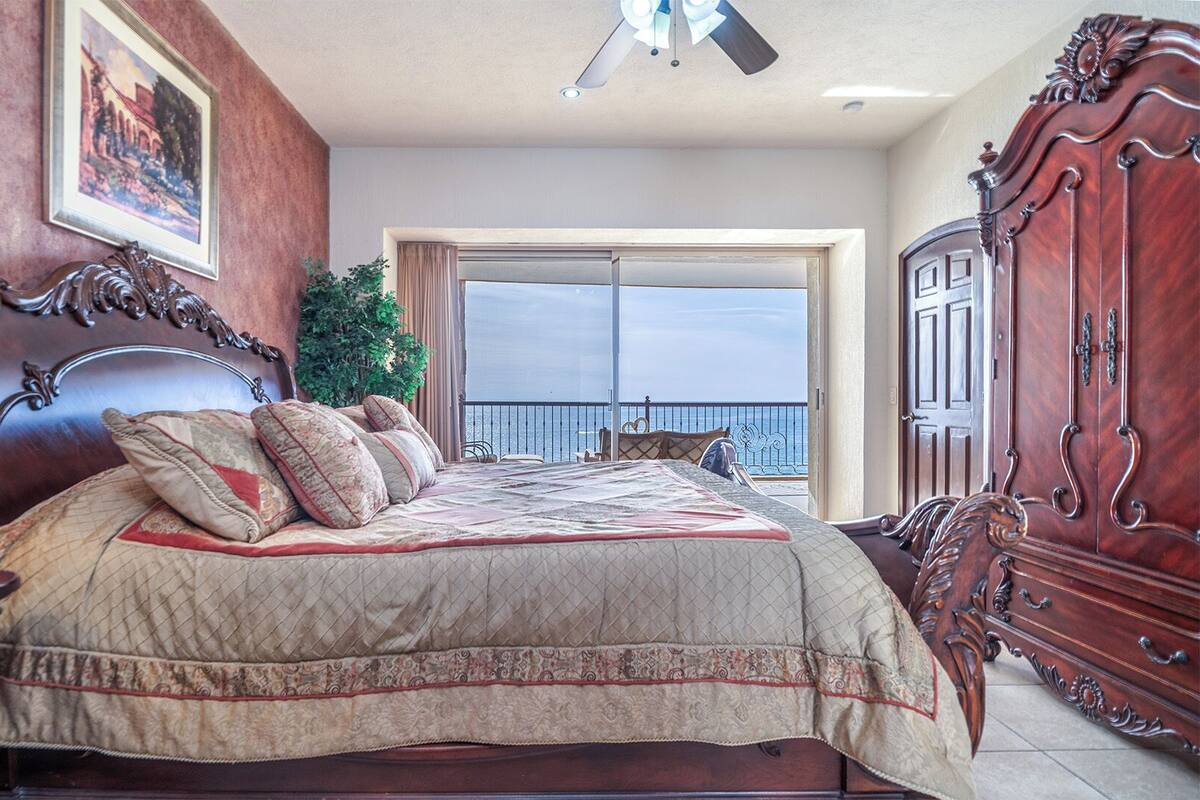 Een slaapkamer met een groot bed, een decoratief houten hoofdbord, een balkon met uitzicht en een plafondventilator.