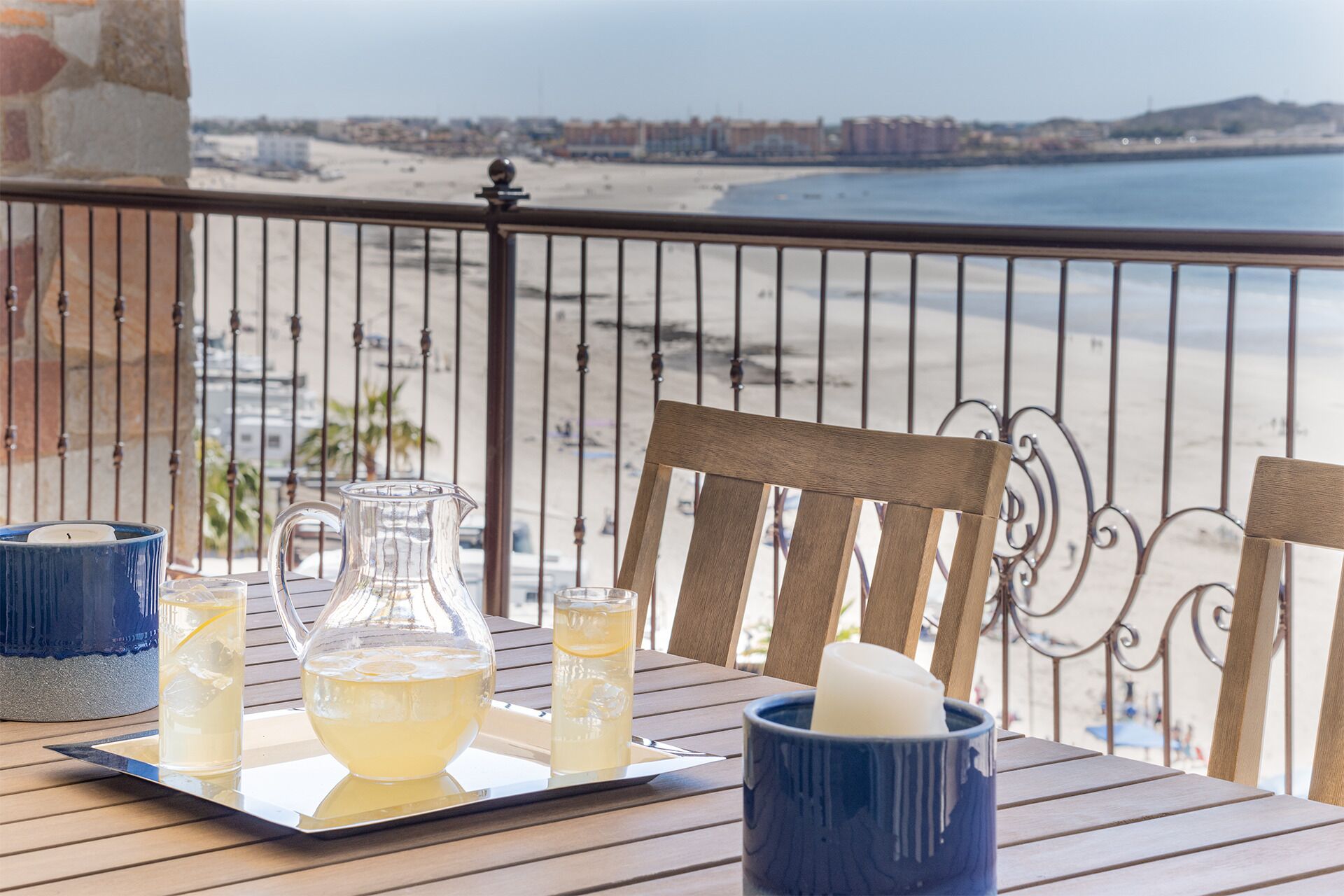 Una mesa con una jarra, vasos y una taza en un balcón con vistas a una playa.