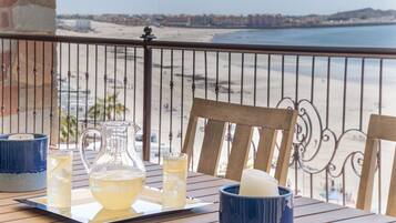 Una mesa con una jarra, vasos y una taza en un balcón con vistas a una playa.