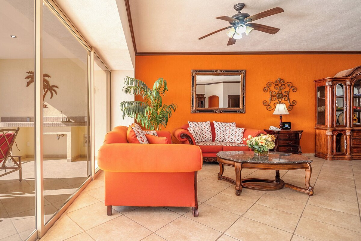 Ein geräumiges Wohnzimmer mit einer großen orangefarbenen Couch, einem Holztisch und einer Essecke mit roten Stühlen. An den Wänden sind ein Deckenventilator und ein großer Spiegel zu sehen.