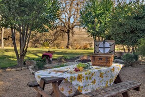 Ein Picknick mit Tisch, der mit einer Blumen-Tischdecke bedeckt ist, einem Korb mit einer Uhr und einem Picknicktisch in einer grünen, offenen Umgebung.