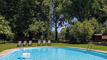 A swimming pool with a clear blue water surface, surrounded by a grassy area with several lounge chairs and a large tree in the background.