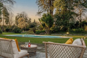A poolside area with wicker furniture and a table, surrounded by lush greenery and trees.