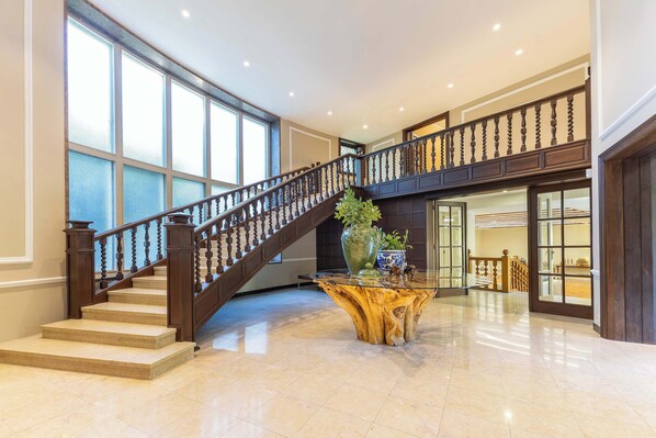 A spacious lobby with a grand staircase, large windows, and a central wooden table.