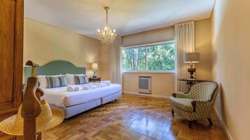 A hotel room with a bed, a chair, a table, a window with curtains, a chandelier, and a wall-mounted air conditioner.