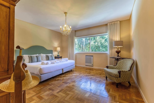 A hotel room with a bed, a chair, a table, a window with curtains, a chandelier, and a wall-mounted air conditioner.