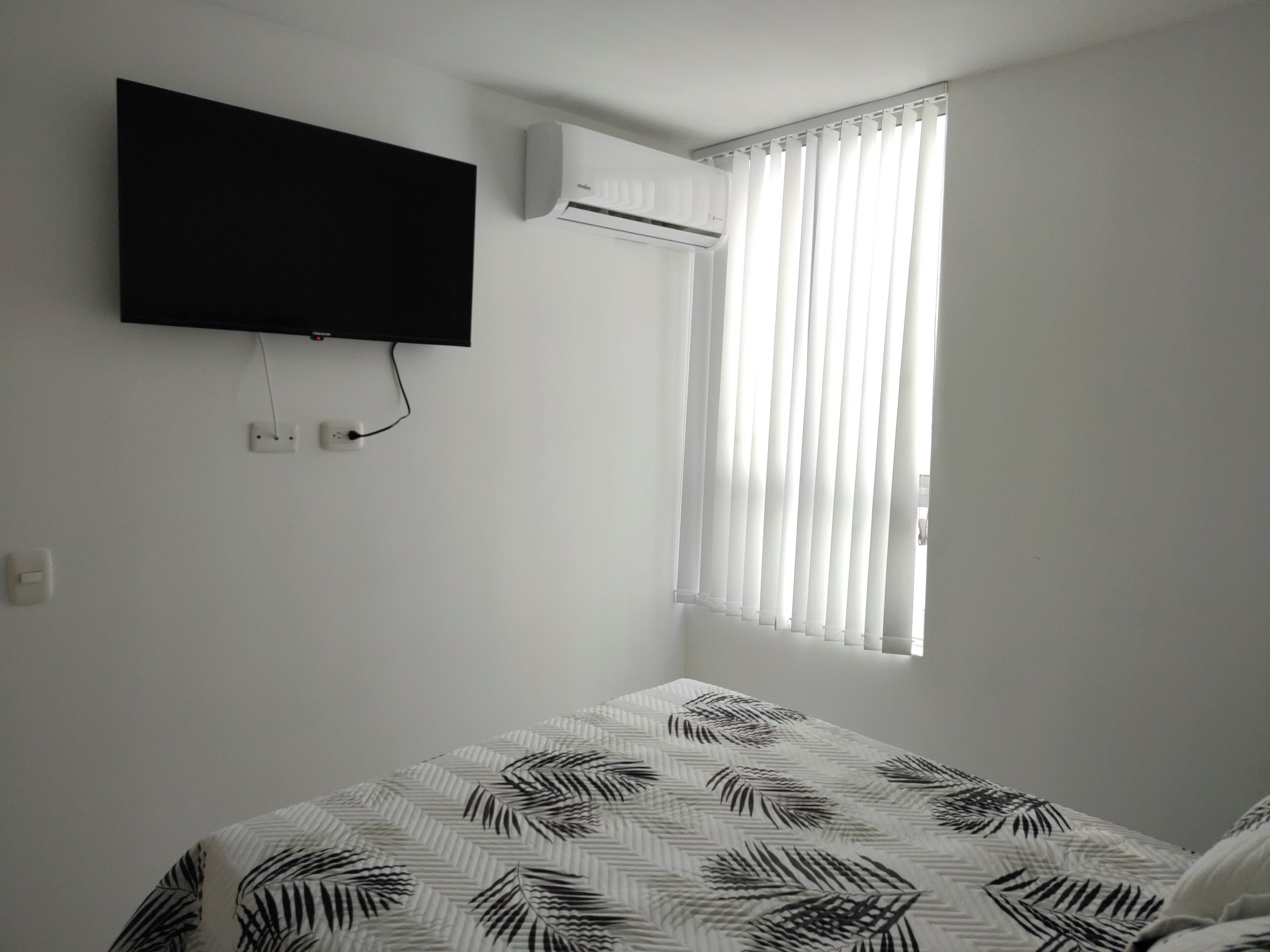A bedroom with a bed, a mounted TV, an air conditioning unit, and a window with vertical blinds.
