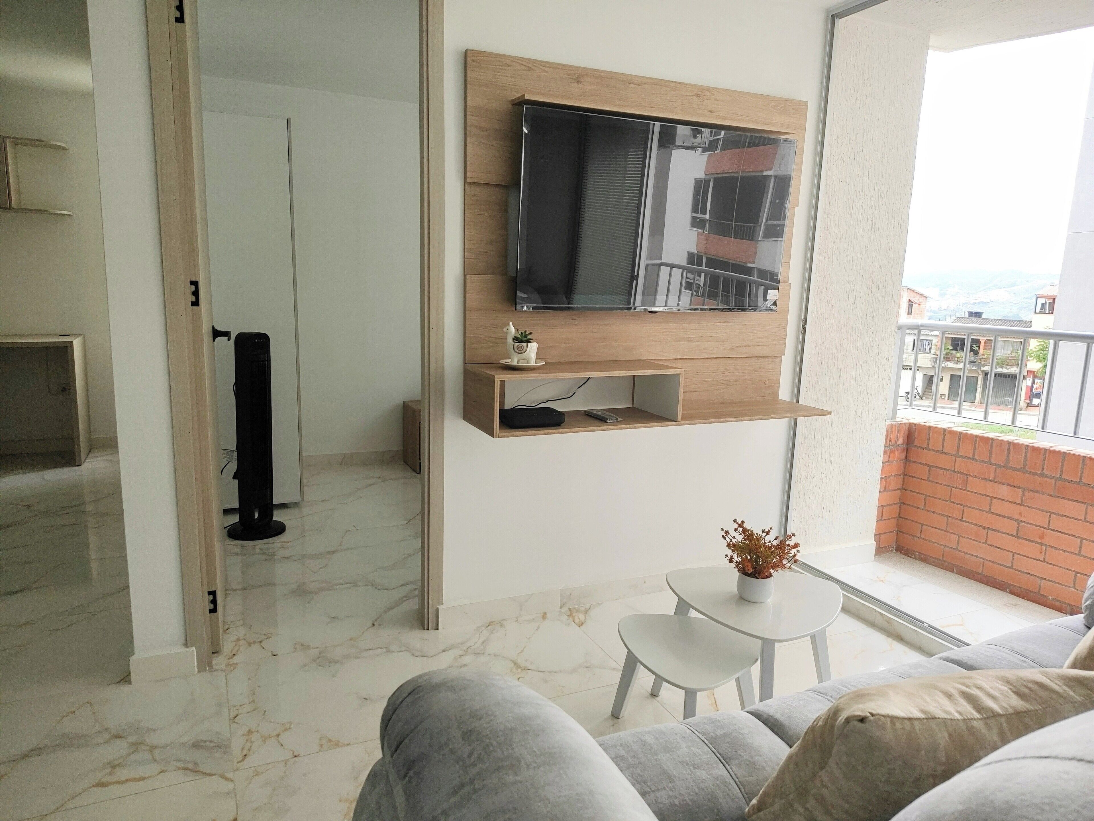 Una sala de estar moderna con un televisor de pantalla plana montado en un estante de madera, una mesita con una planta en maceta y un sofá gris.