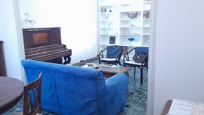 A living room with a blue sofa, a wooden coffee table, a piano, and a white bookshelf. - Ravenna Suite: apartment close to the center and major monuments! (Ravenna)