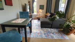 A living room with a sofa, a dining table, and chairs. There is a window with curtains, a lamp, and a plant. - Spruce Suite - Beach and Vineyard steps away! (Cobble Hill)
