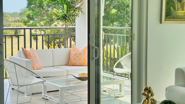 Uma sala de estar moderna com uma porta de vidro que dá para uma varanda. O espaço possui um sofá branco, uma mesa de centro de vidro e uma poltrona branca. Há uma pequena mesa com objetos decorativos e um quadro na parede.