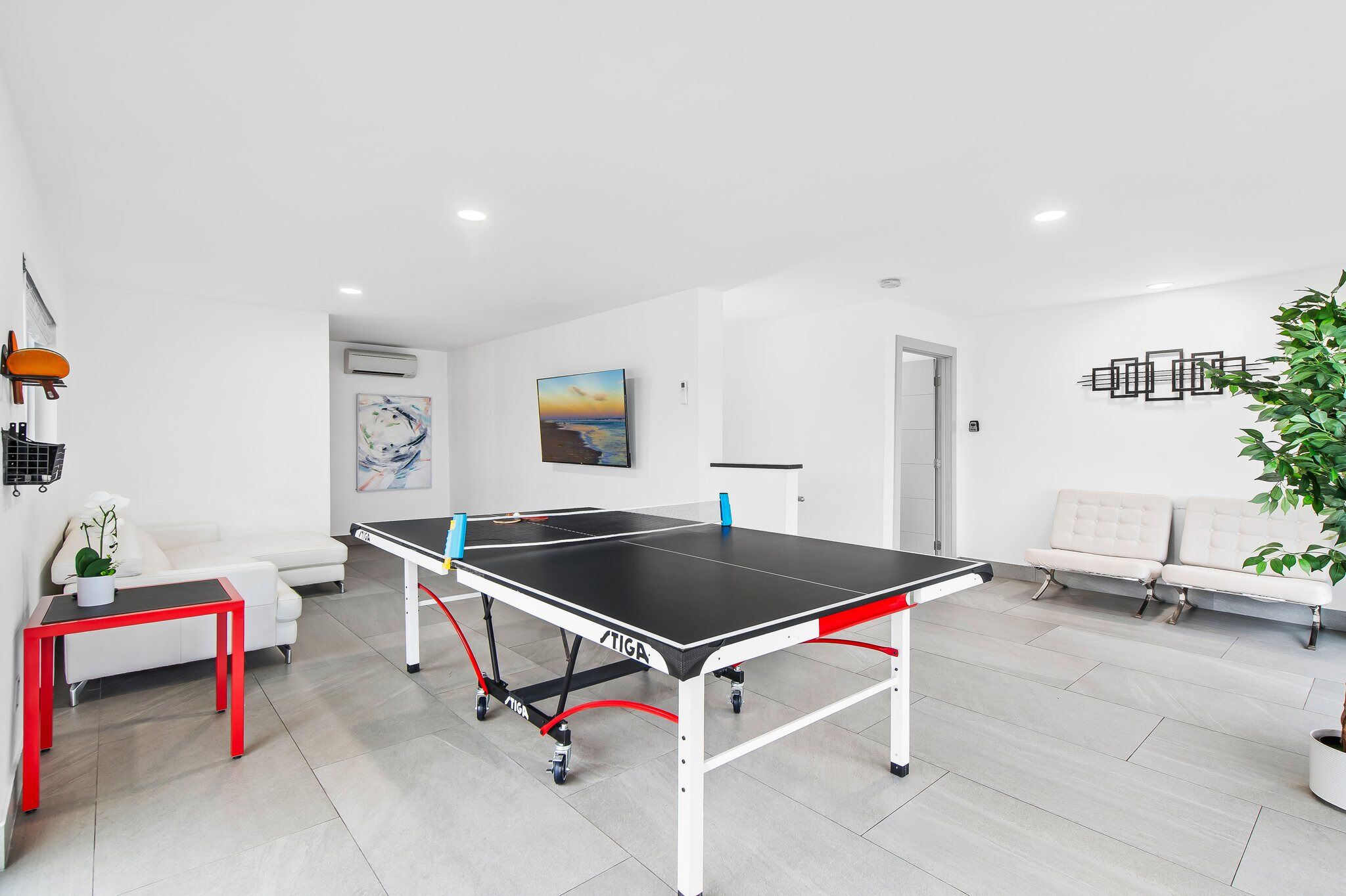 A ping pong table in a modern, well-lit room with a white couch, a small table with a plant, and a painting on the wall.