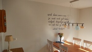 A dining area with a wooden table, chairs, a vase with flowers, and a wall with a quote in German.