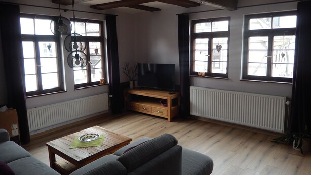 A living room with a sofa, a coffee table, a TV on a wooden stand, and large windows with curtains.