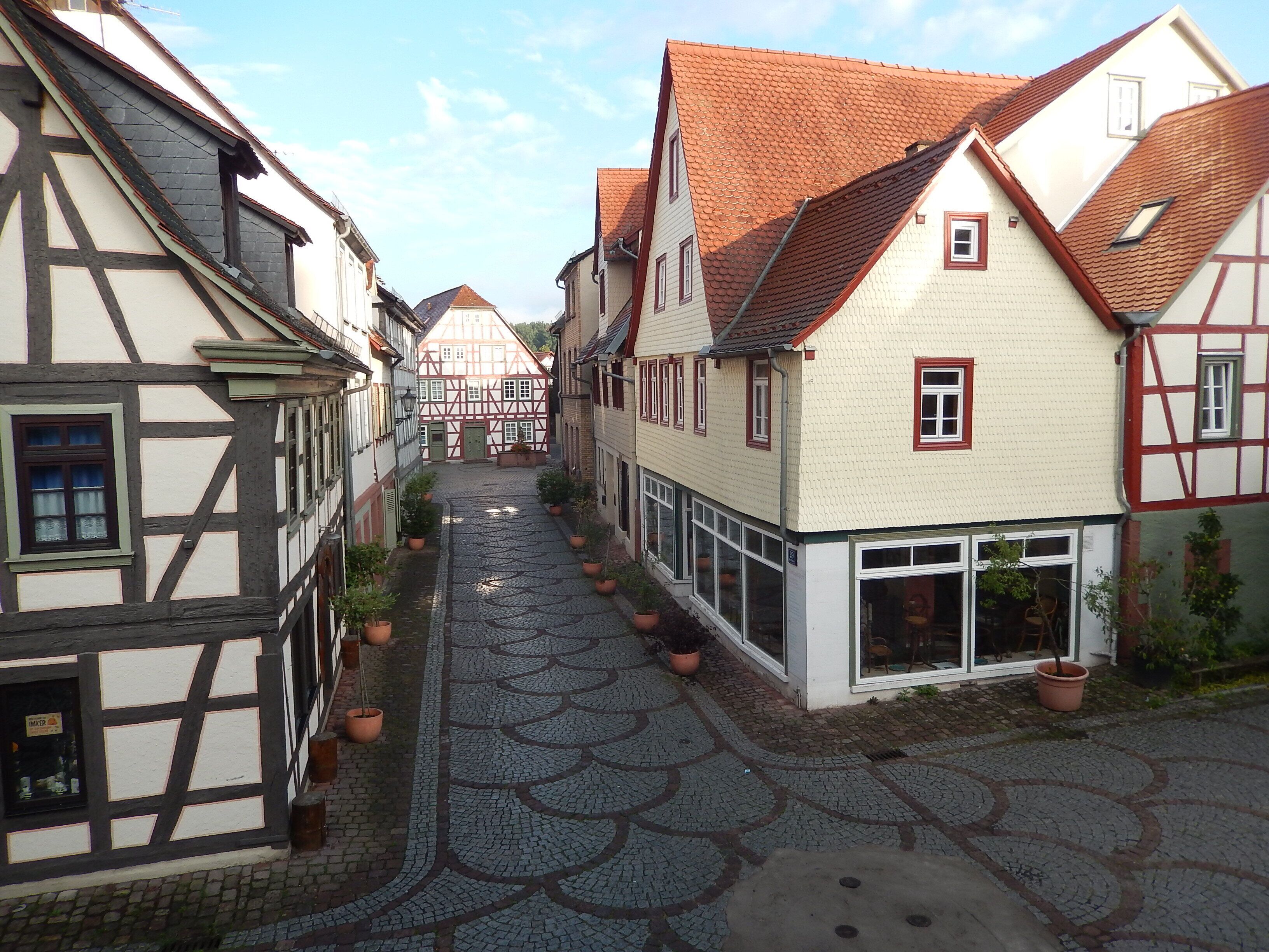 Eine enge Kopfsteinpflasterstraße gesäumt von traditionellen Fachwerkhäusern, mit einer Mischung aus weißem und dunklem Fachwerk, und ein klarer Himmel darüber.