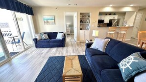 Una sala de estar con un sofá azul oscuro, un banco de madera y una alfombra. Se observa una cocina al fondo con armarios blancos y una zona de comedor con sillas.