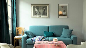 Una sala de estar con un sofá verde azulado, una mesita con mantel a cuadros y dos cuadros en la pared.