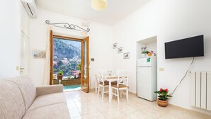A living room with a sofa, a dining table with chairs, a TV mounted on the wall, and a door leading to a balcony with a view of a town and mountains. - Rosa House (Positano)