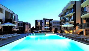 Piscina com espreguiçadeiras e guarda-sóis, cercada por edifícios com varandas e aparelhos de ar condicionado.