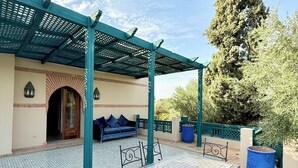 A covered outdoor seating area with a tiled floor, a metal table with chairs, a blue sofa, and a large tree in the background. - VILLA 34 - 4 Masters, Moroccan Charm & Modern Comfort with Private Pool (Marrakech)