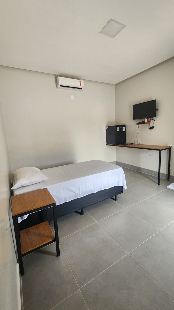 A single bed with a white mattress and a wooden headboard, a small bedside table, a wall-mounted air conditioner, a flat-screen TV on a stand, and a desk with a chair. - Friper Hotel (Jaraguá)