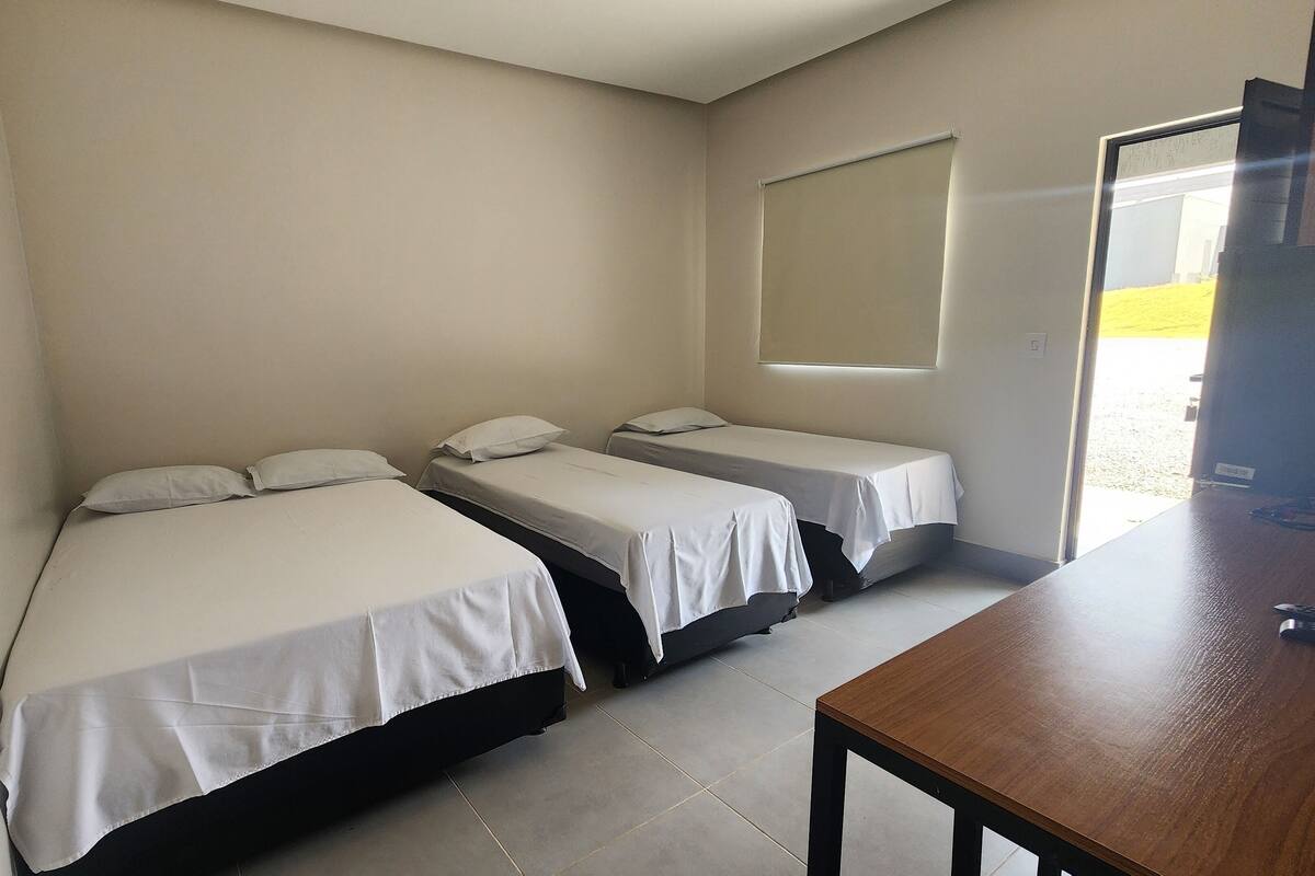 A room with three single beds, a wooden table, and a window with blinds.