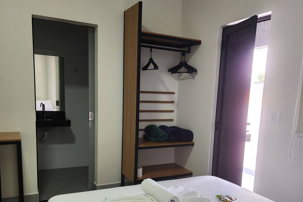 A hotel room with a bed, a bedside table, a mirror, a wardrobe with hanging space, and a door leading to another room.