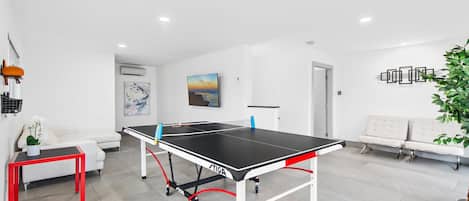 A ping pong table in a modern room with a white sofa, a small table with a plant, and a painting on the wall.