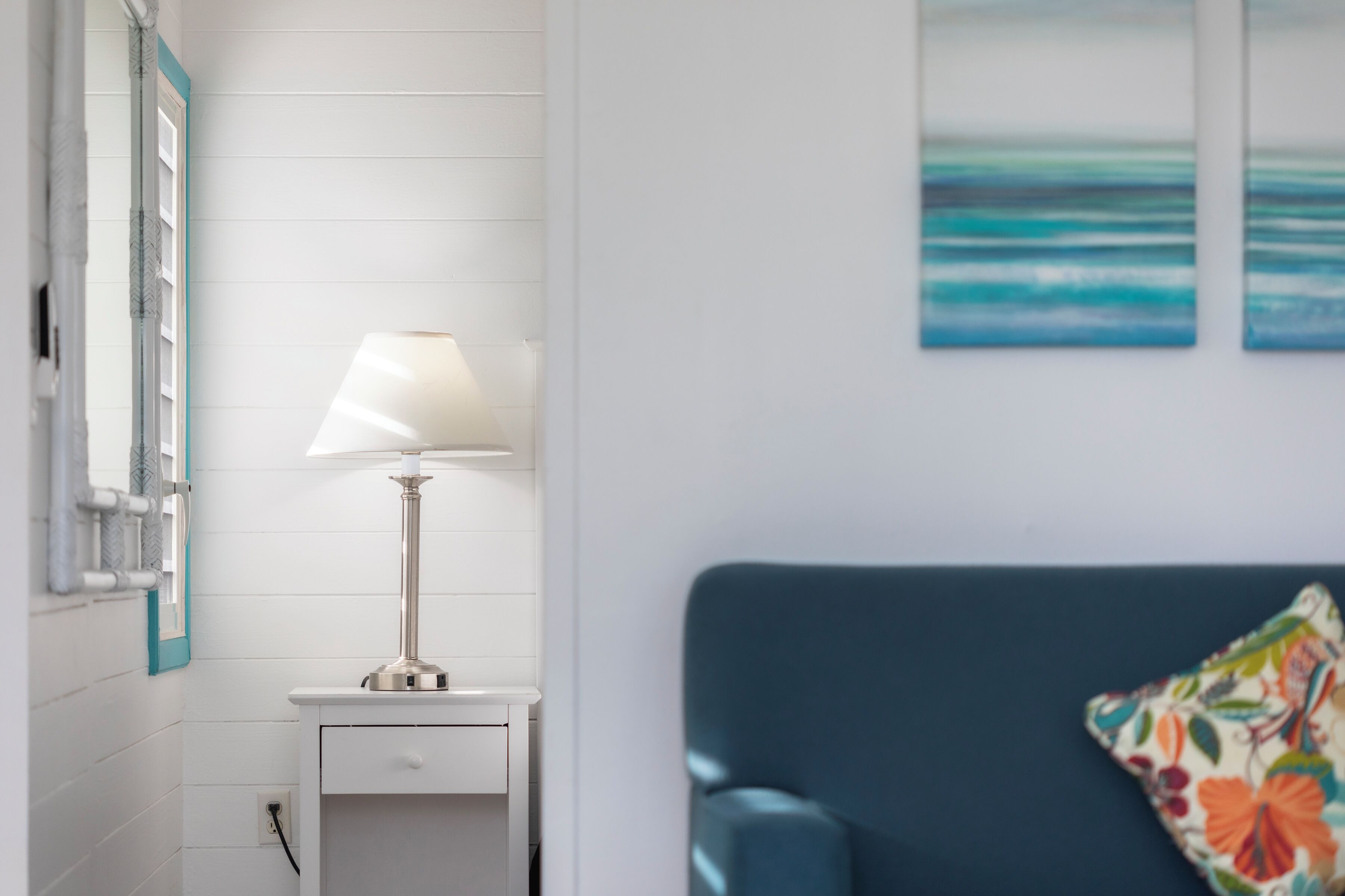 Una habitación con pared blanca, un sofá azul, una mesita de noche blanca con lámpara y una ventana con vista al océano.