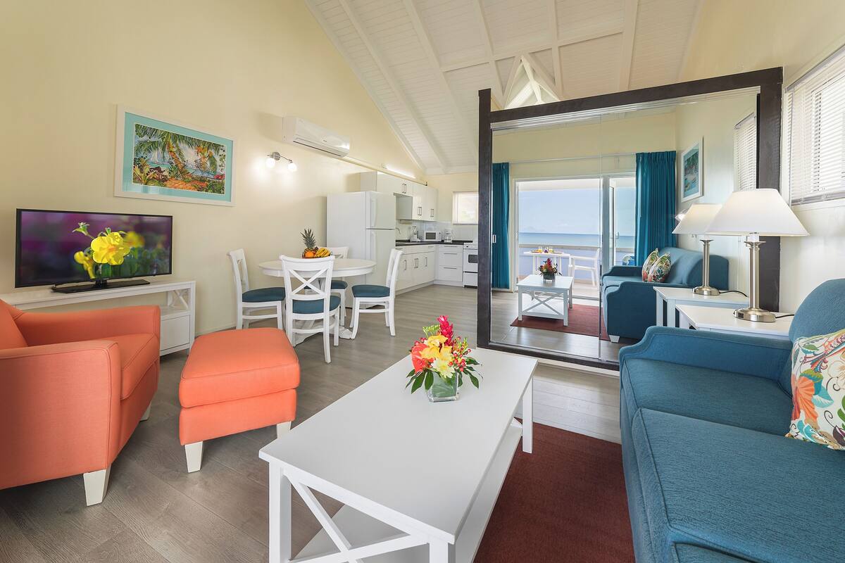 Una sala moderna con sofá, sillón y mesa de centro. Incluye televisión, mesa de comedor con sillas y una zona de cocina con refrigerador y microondas.