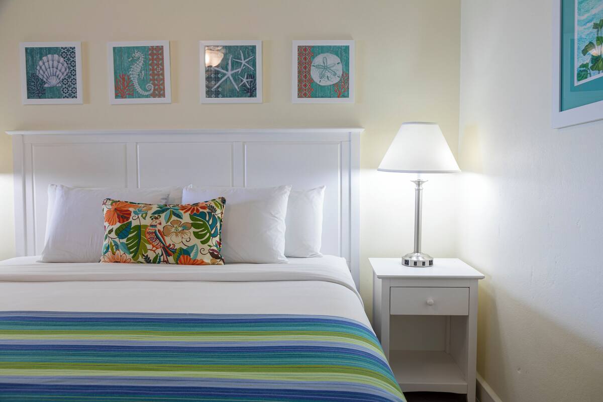 Una cama bien tendida con un cojín colorido, una mesita de noche blanca con una lámpara y tres cuadros enmarcados en la pared.