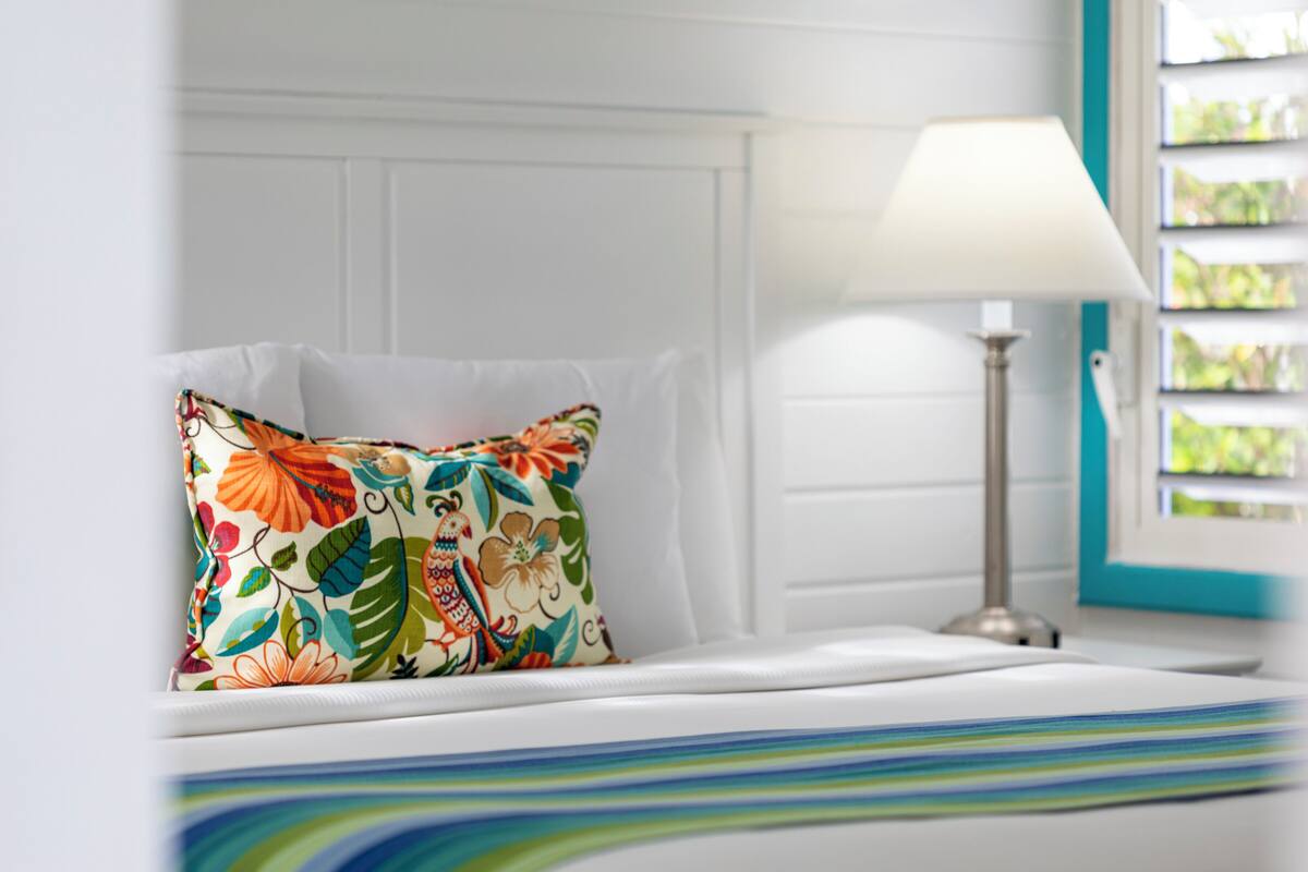 Una cama bien hecha con un cojín floral colorido, un cabecero blanco y una lámpara de noche.