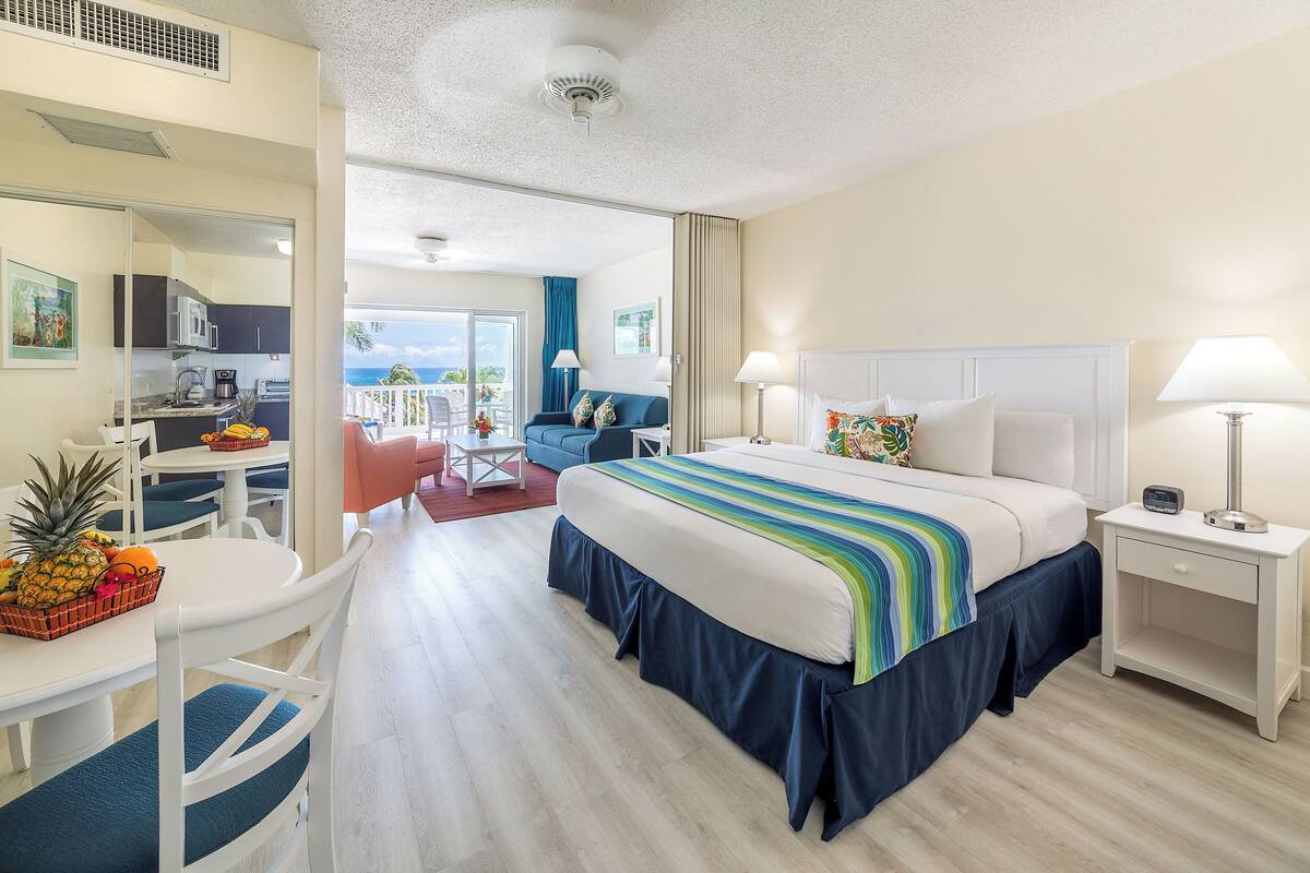 Una habitación de hotel con cama, mesitas de noche, un escritorio con sillas, una kitchenette y un balcón con vistas.