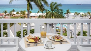 A balcony table with breakfast items, a glass of orange juice, and a view of the ocean and palm trees. - La Vista Hotel (Simpson Bay)