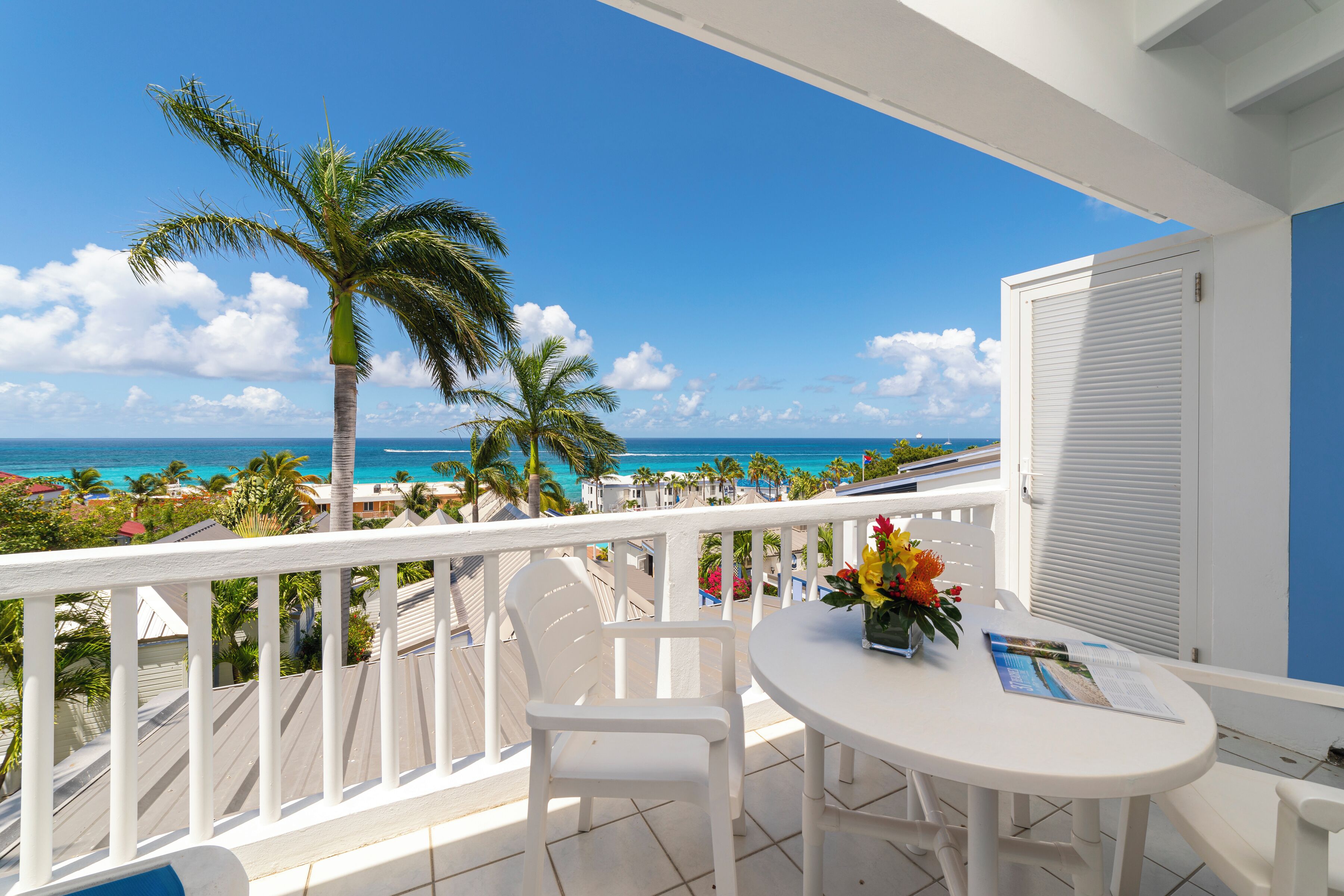 Un balcon avec une balustrade blanche, une table avec des chaises et une vue sur l’océan et les palmiers.