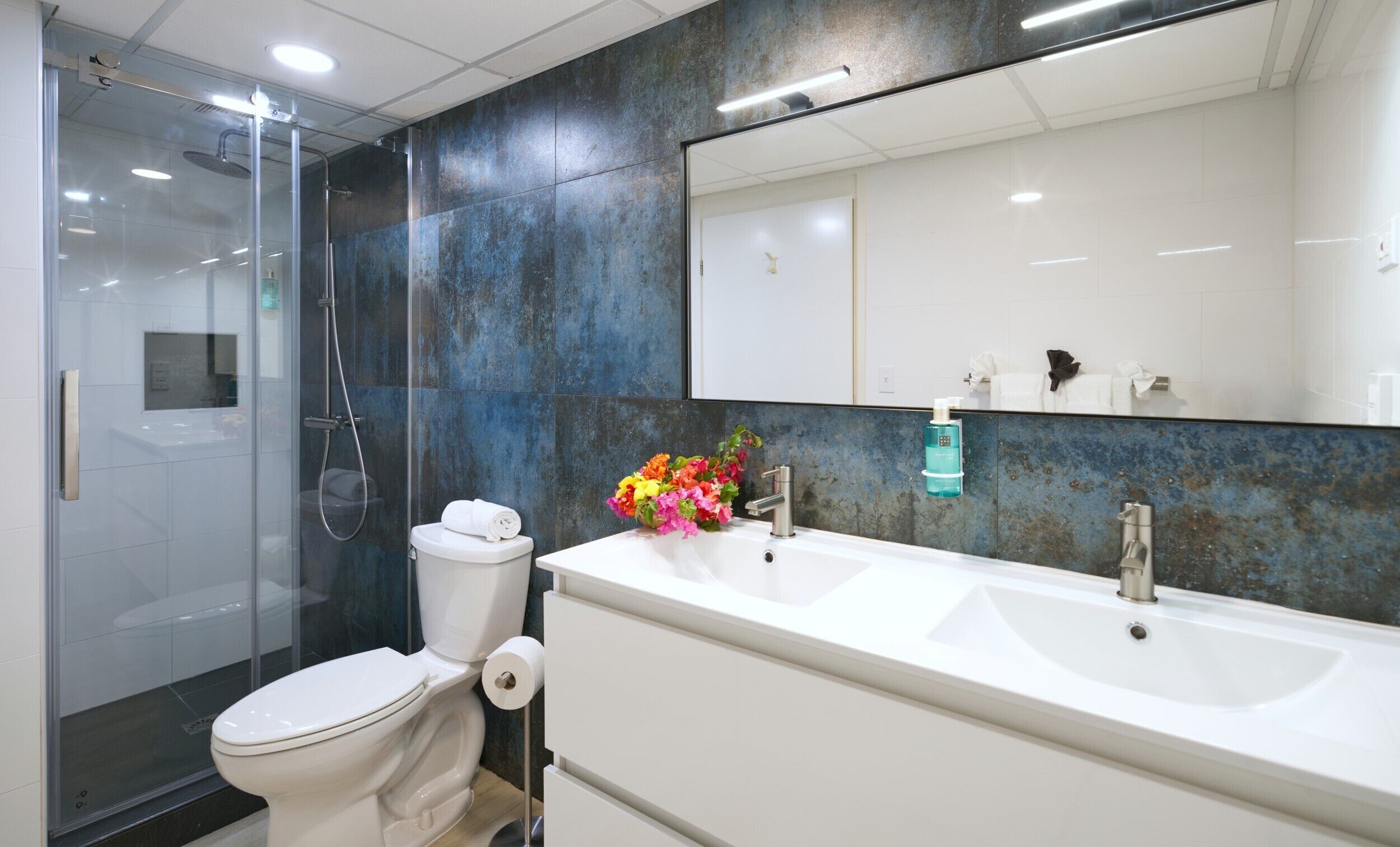 A modern bathroom with a glass-enclosed shower, a white toilet, a large sink with a mirror above it, and a decorative flower arrangement.