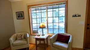 A room with two armchairs, a small wooden table, a lamp, and a window with a view of trees and a building.