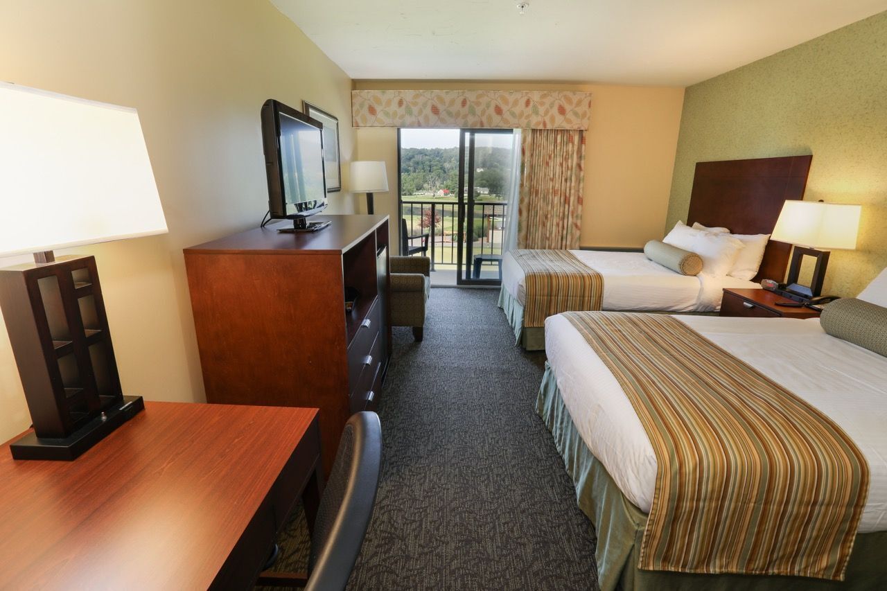 A hotel room with two beds, a desk with a television, a chair, and a view of the outdoors through a sliding door.
