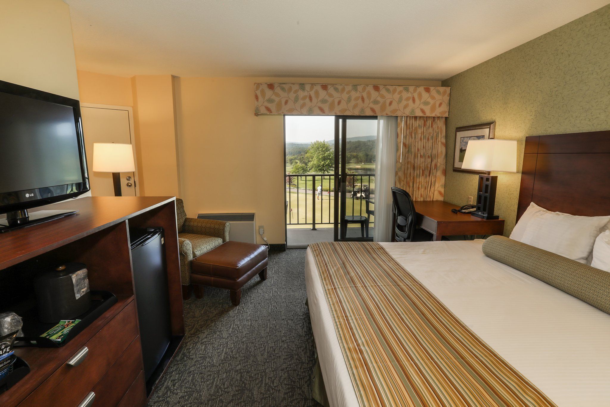 A hotel room with a bed, a desk, a television, a refrigerator, a chair, and a view of the outdoors.