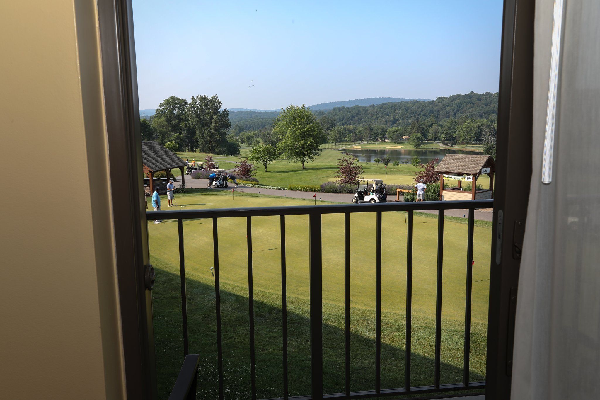 A view from a balcony overlooking a golf course with a pond, trees, and distant hills.