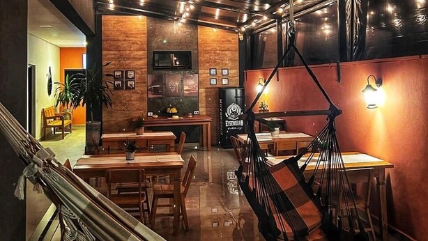 A restaurant interior with wooden tables, chairs, a hanging hammock, and wall-mounted lights. - Pousada Lobo Guará (São João Batista do Glória)