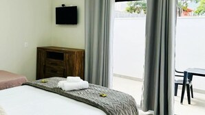 A hotel room with a bed, a wooden nightstand, a TV mounted on the wall, and a view of the street through large windows. - Pousada Lobo Guará (São João Batista do Glória)