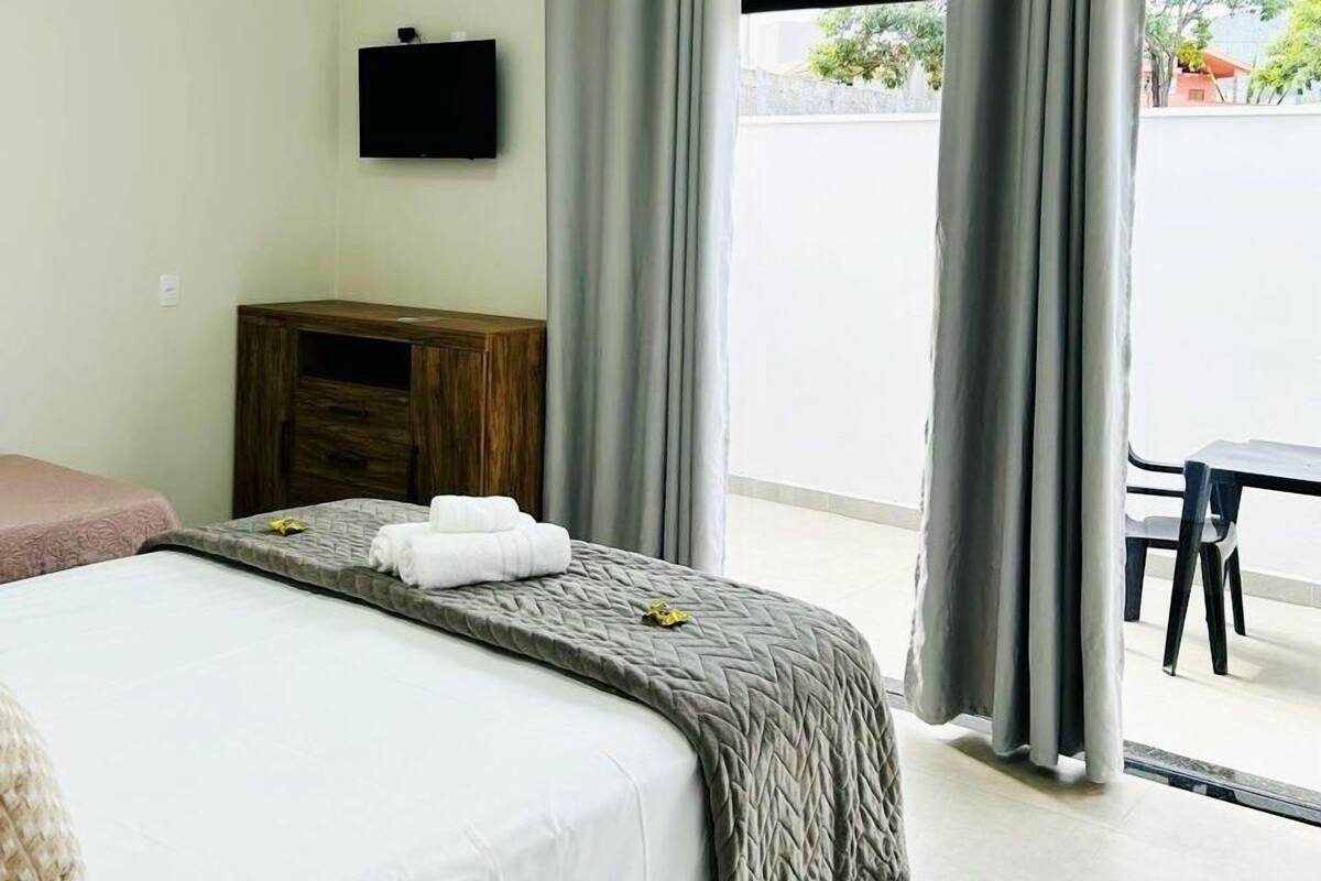 Habitación de hotel con una cama, una mesita de noche de madera, un televisor montado en la pared y vistas a la calle a través de amplias ventanas.